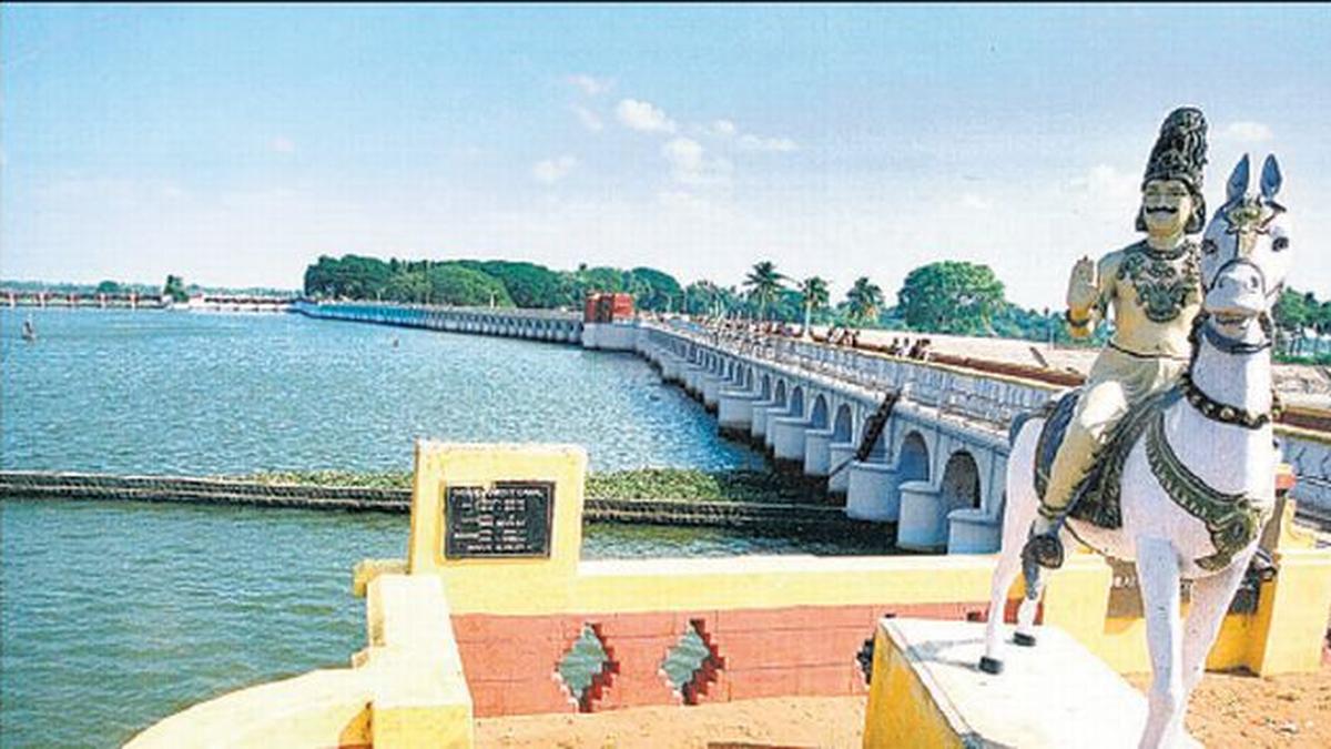 Photograph of the historic Kallanai (Grand Anicut) dam across the Kaveri River in Tamil Nadu, showing the long stone barrage with arched openings, calm blue water, and a statue of Karikala Chola on horseback holding a trident in the foreground.
