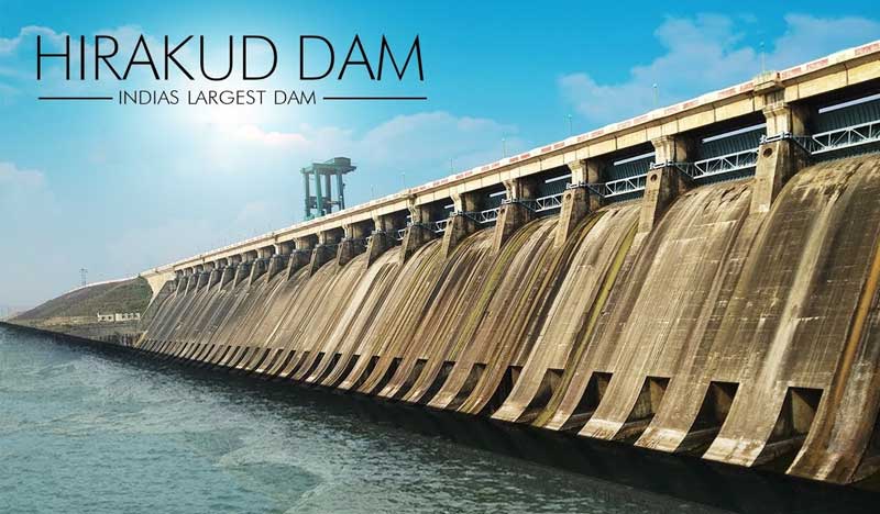 Front view of Hirakud Dam showing a long concrete spillway structure with multiple gates, water flowing below, and the title “Hirakud Dam – India’s Largest Dam” displayed in the sky.