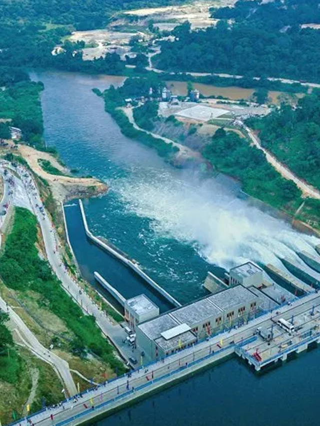 Aerial view of Hirakud Dam and its hydroelectric power station, with water being released into the river below, surrounded by green hills and infrastructure buildings.